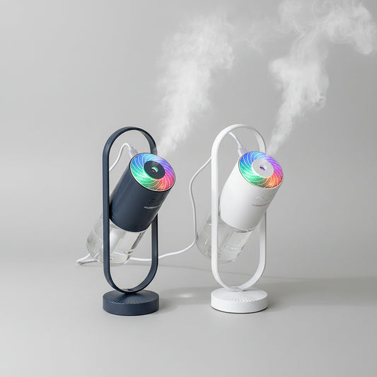 Two portable mist humidifiers with cylindrical water tanks and illuminated rainbow LED tops, shown in a styled indoor setting with visible mist and a glass vase in the background.